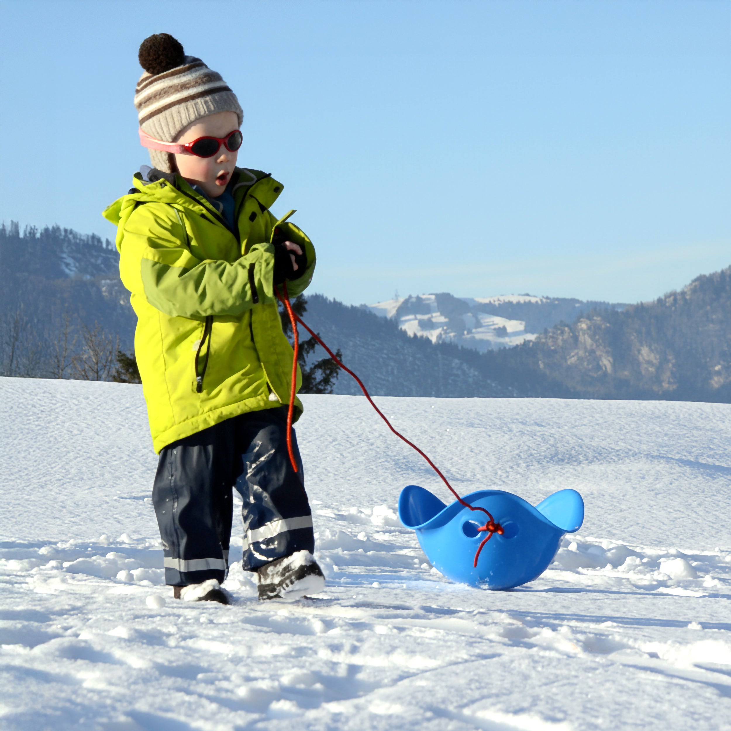 Child in winter clothes pulling a blue Bilibo play shell on a rope like a sled in a snowy landscape with mountains in the background.