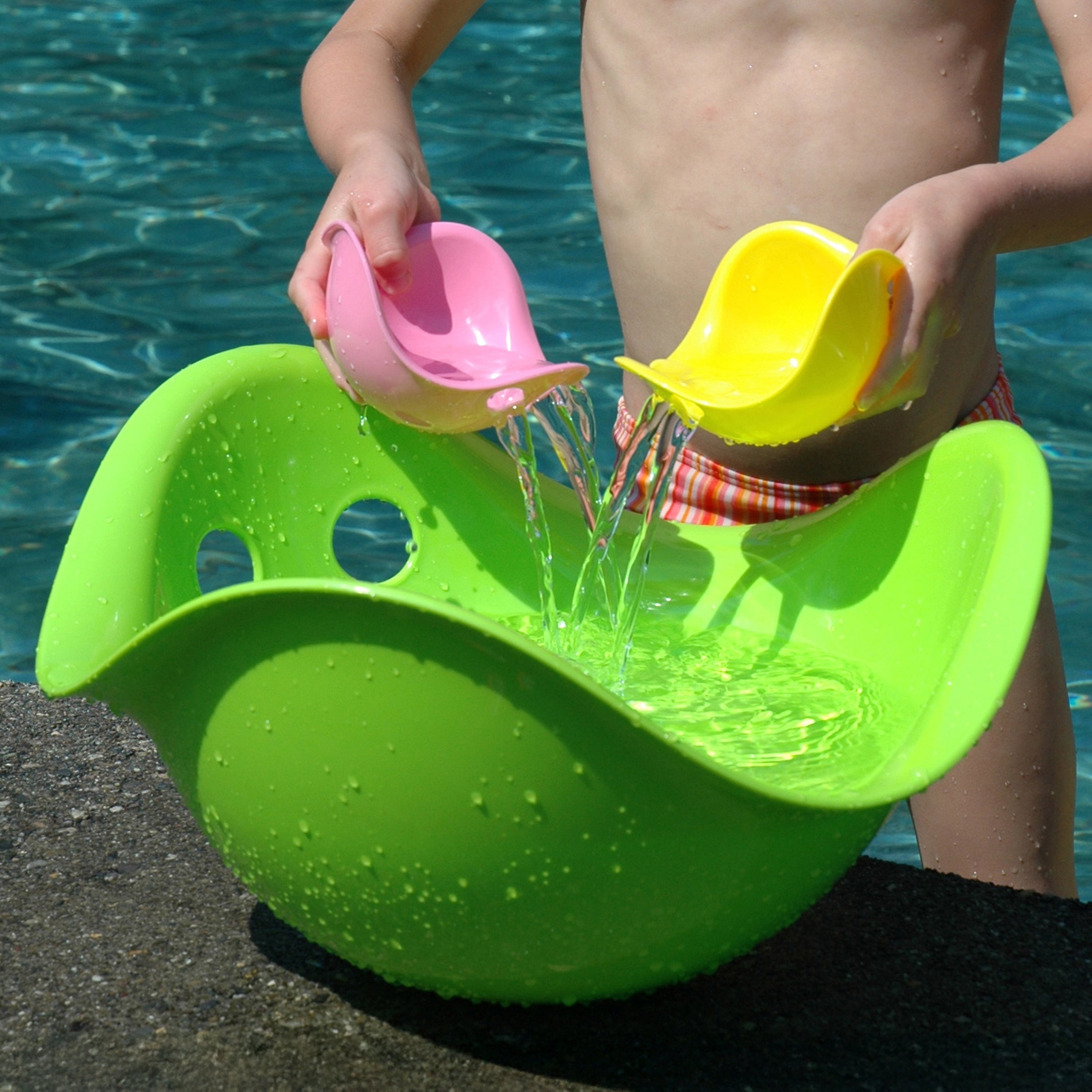 A child pouring water from two Bilibo Mini sensory toys into a large green Bilibo play shell by MOLUK at the pool.