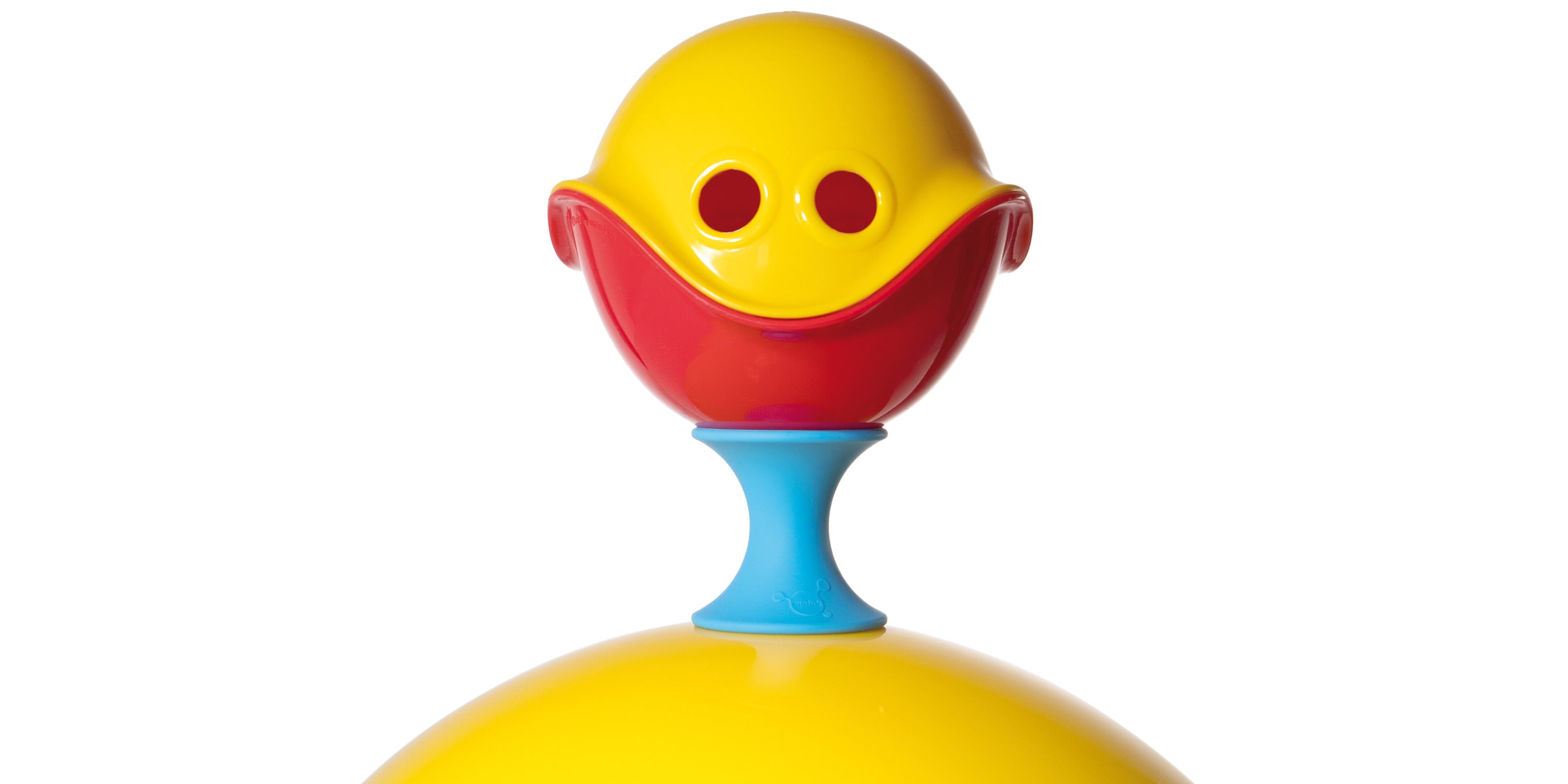 A red and a yellow Bilibo Mini shell are joined to form a smiling sphere nesting on a blue Tulu silicone building block by MOLUK.