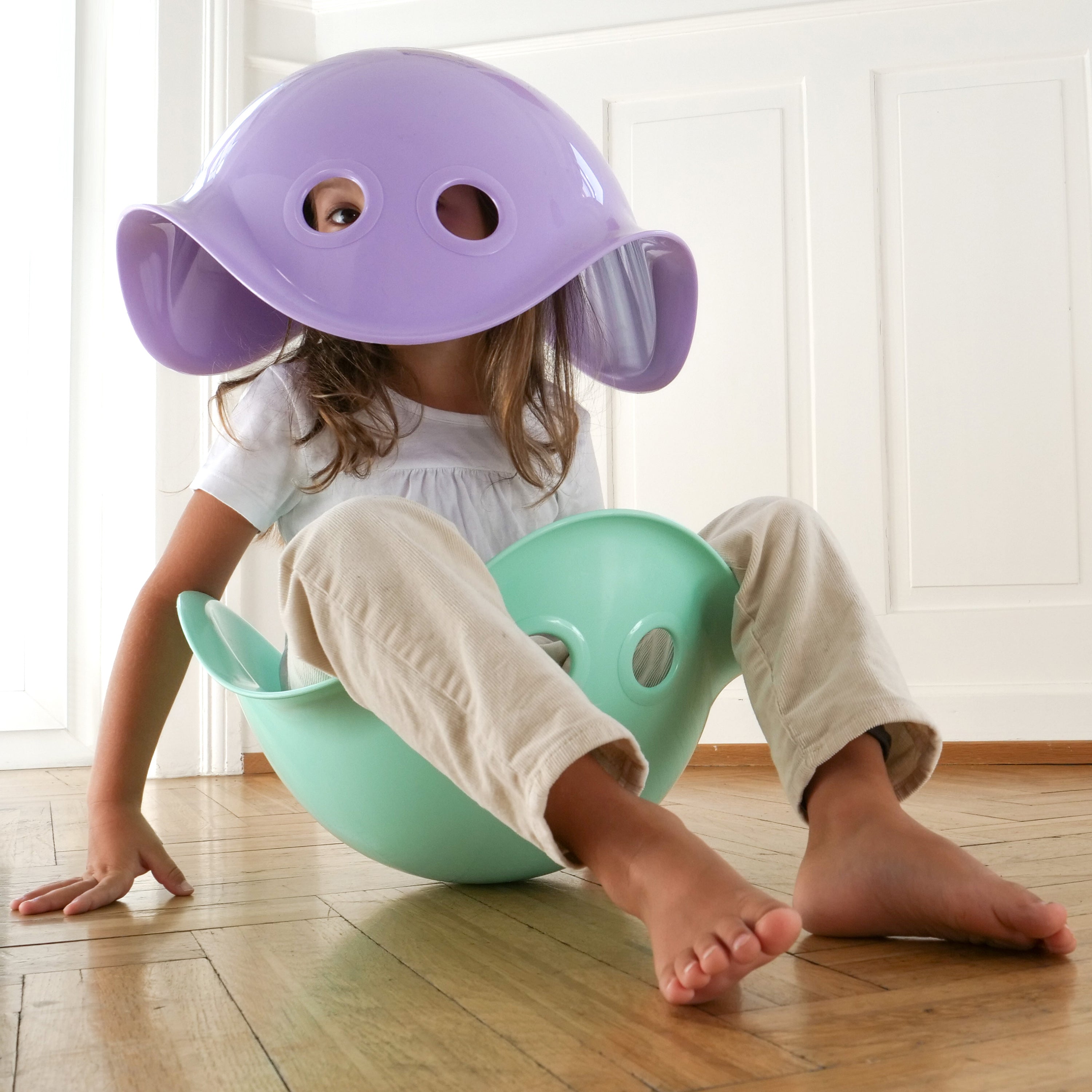 Girl sitting inside a mint Bilibo play shell by MOLUK, with a lilac Bilibo toy like a helmet on her head, peeking through its two holes.