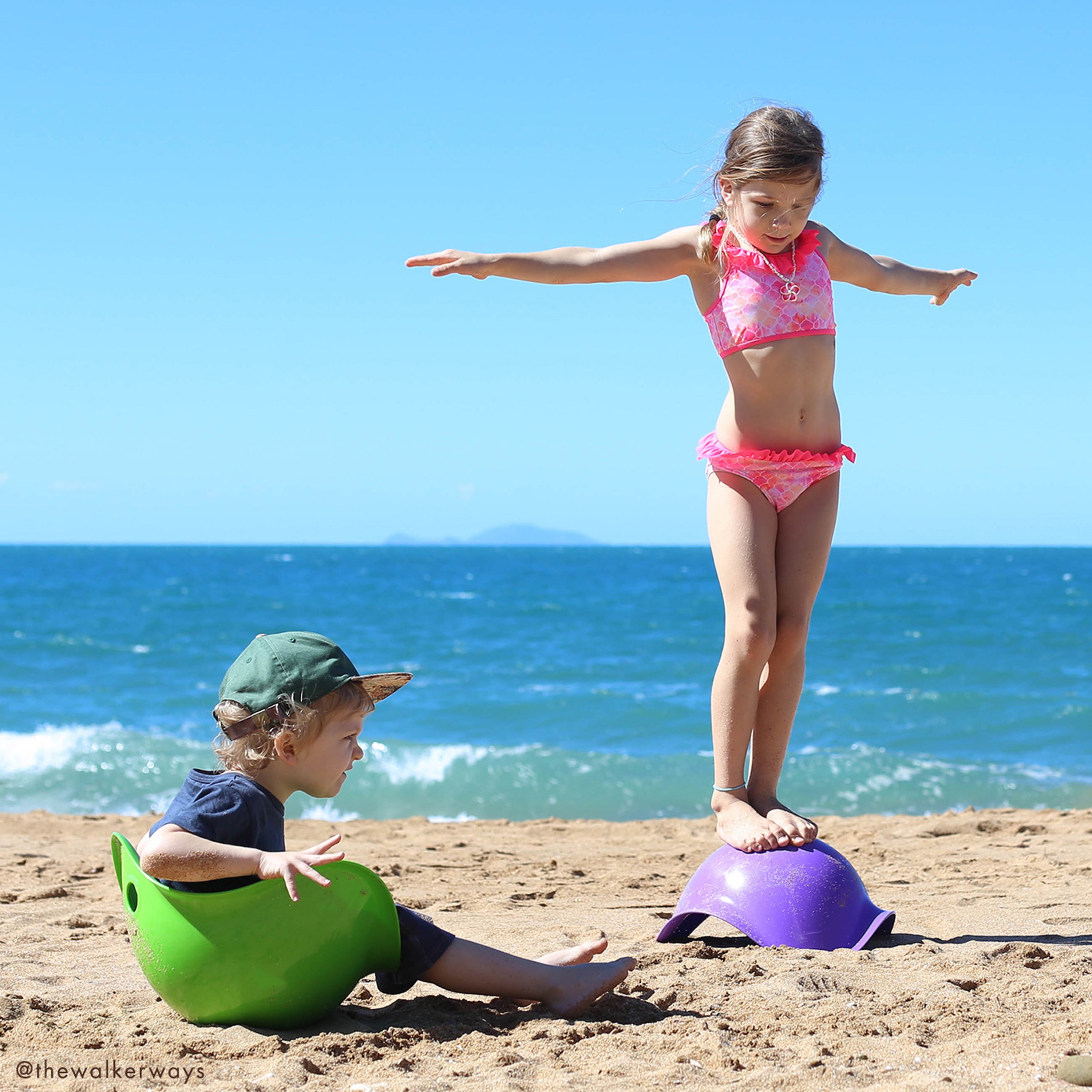 Two children playing on the beach, sitting and balancing on green and purple Bilibo play shells by MOLUK.