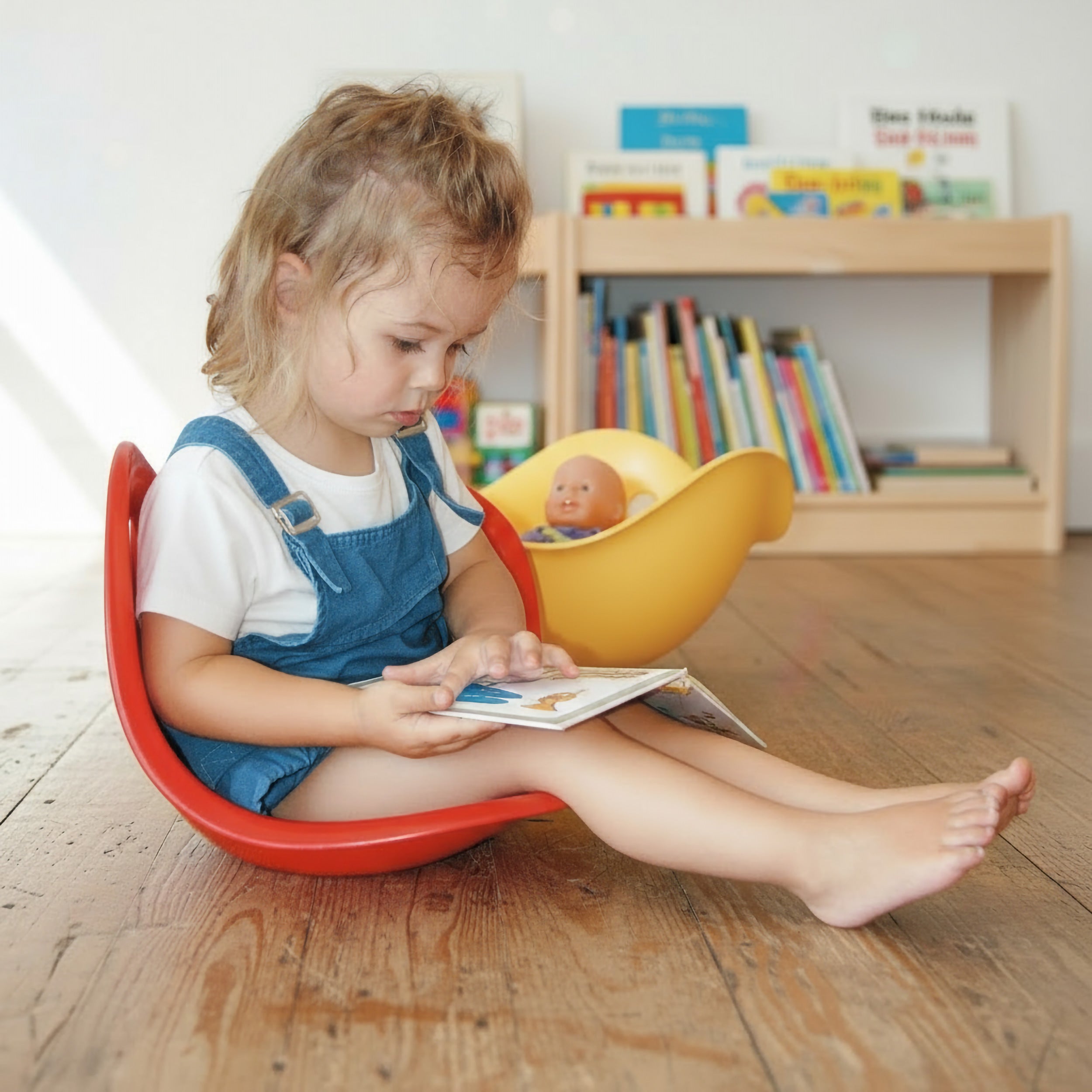 Girl sitting inside a red Bilibo chair reading a children's book, with a yellow Bilibo play shell by MOLUK as a cradle for her doll and a bookshelf in the background.