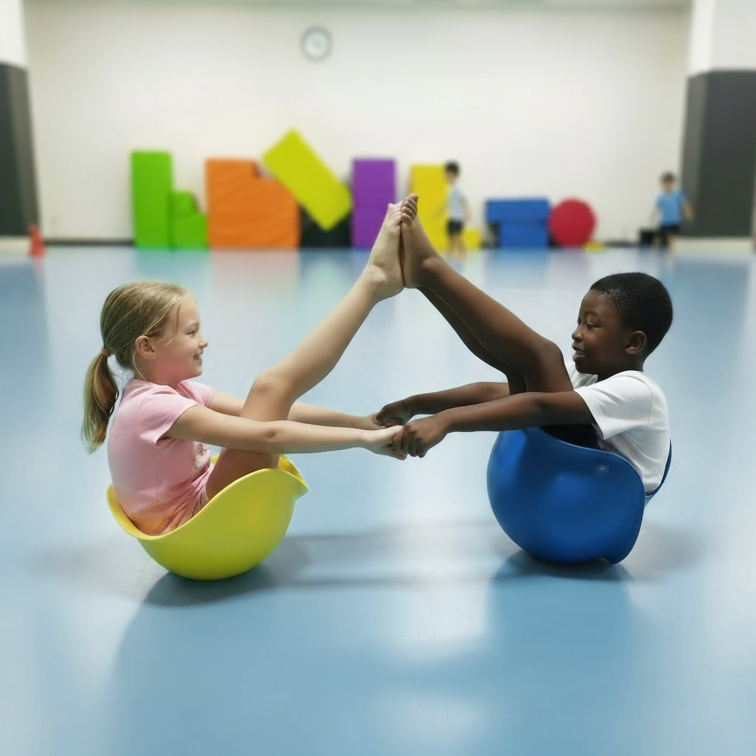 A girl and a bow sitting inside a blue and yellow Bilibo balance and spinning toy by MOLUK, hands and feet touching, in a children's gymnastic class.