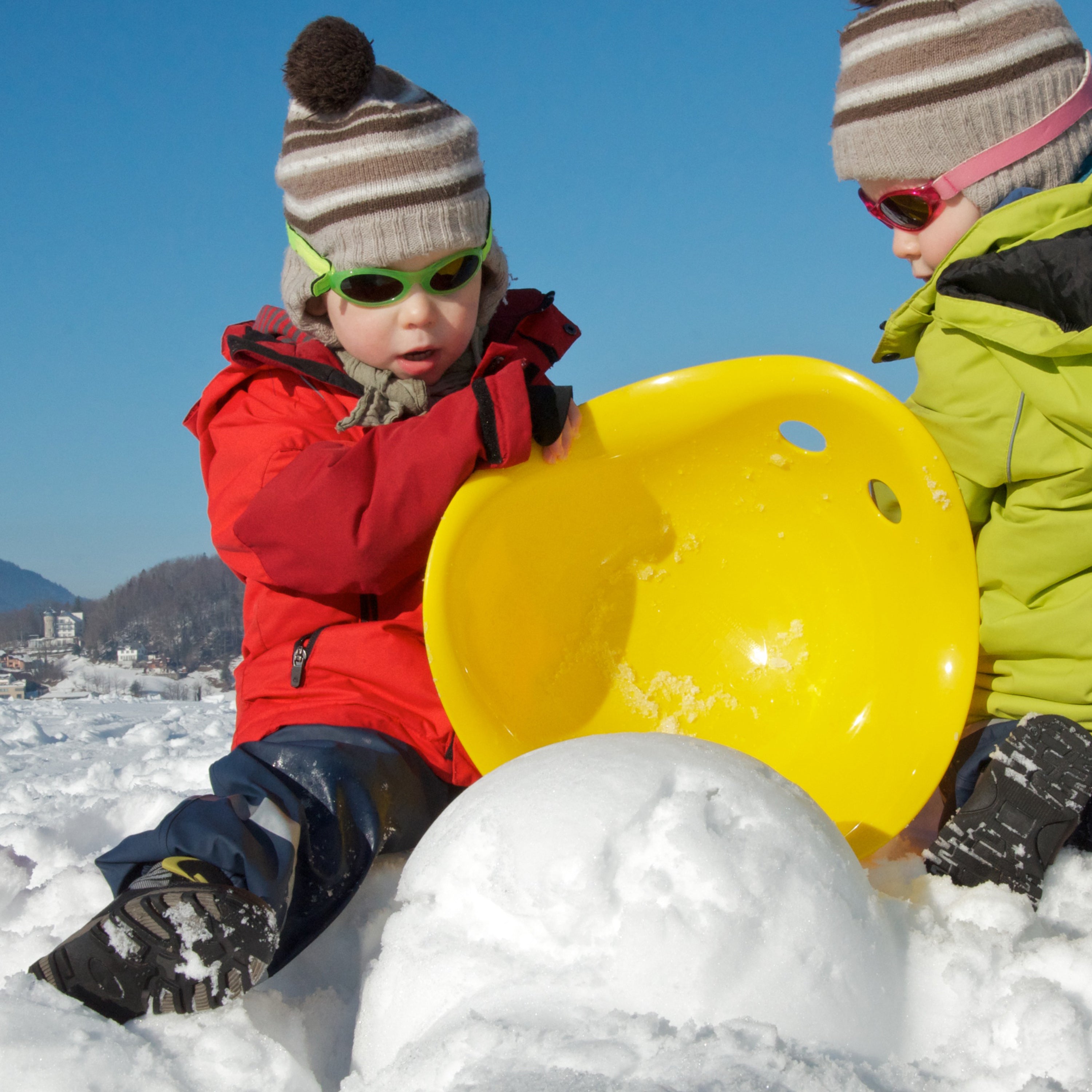 Two children in winter clothing using a yellow Bilibo play shell by MOLUK as a snow mold.
