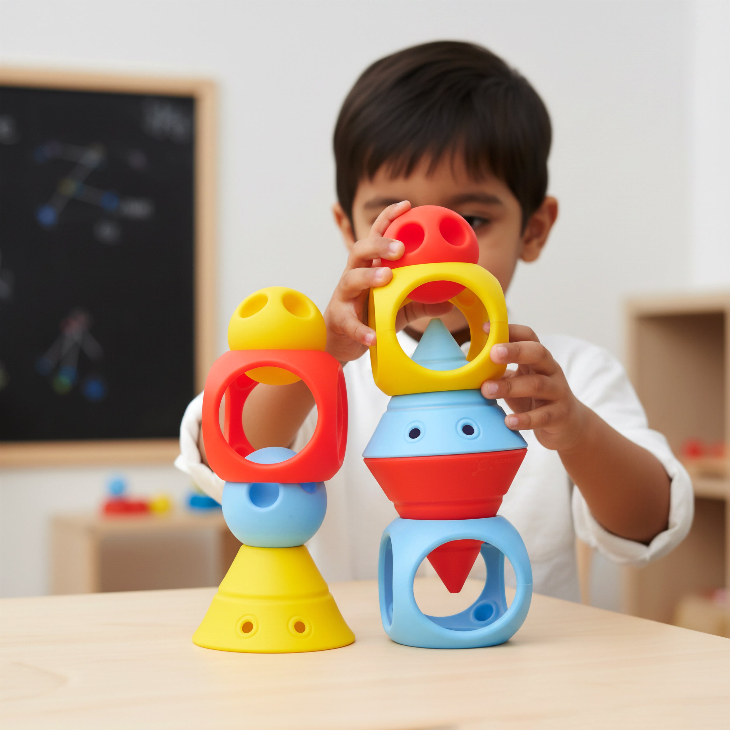 Boy in an elementary school playing with the BUILDING GENIUS set by MOLUK, stacking Hix cones, Mox balls, and Oibo blocks in bright primary colors into imaginative towers.