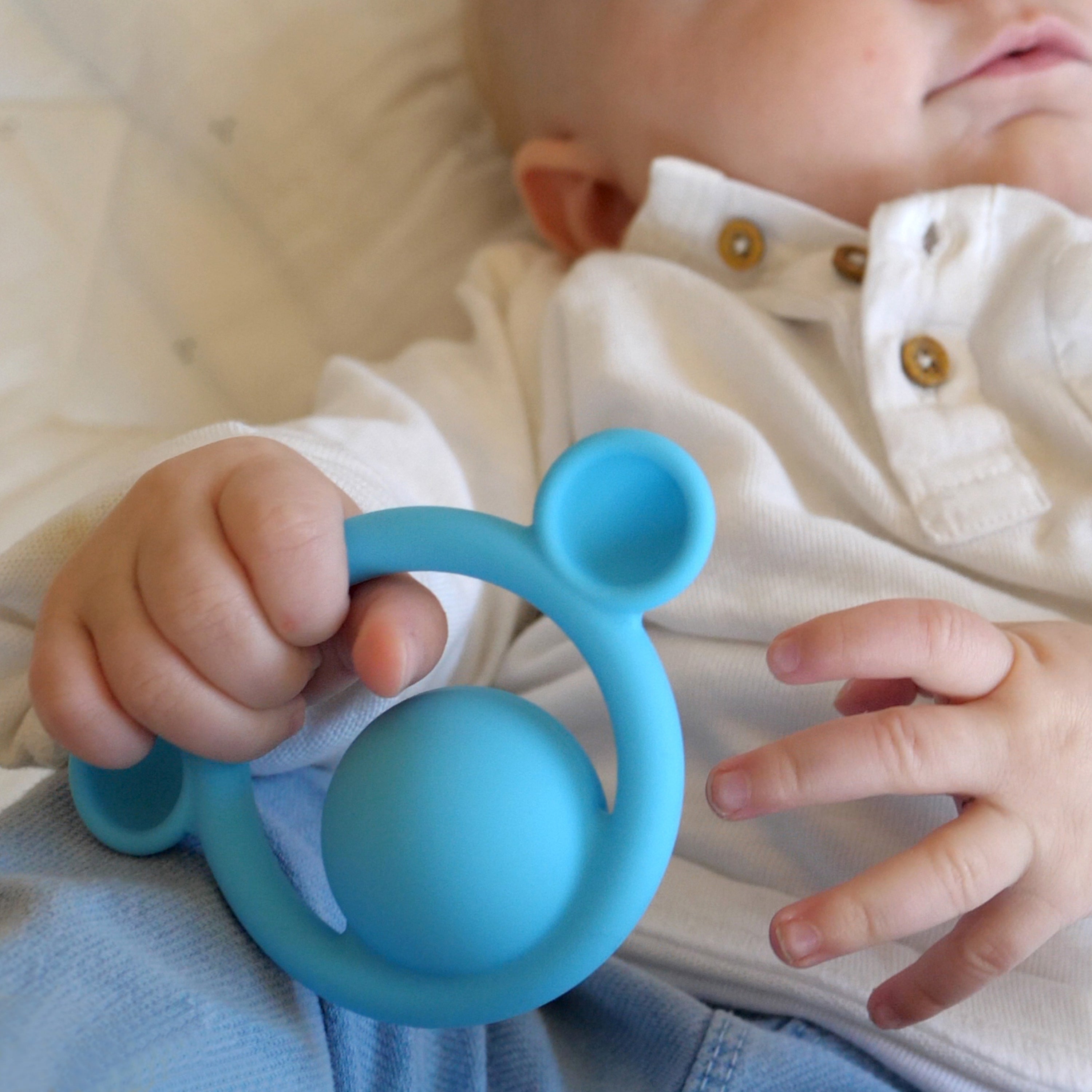 Baby holding a blue Nigi Nagi Nogi silicone teething ring by MOLUK, a tactile toy for grasping and sensory stimulation.