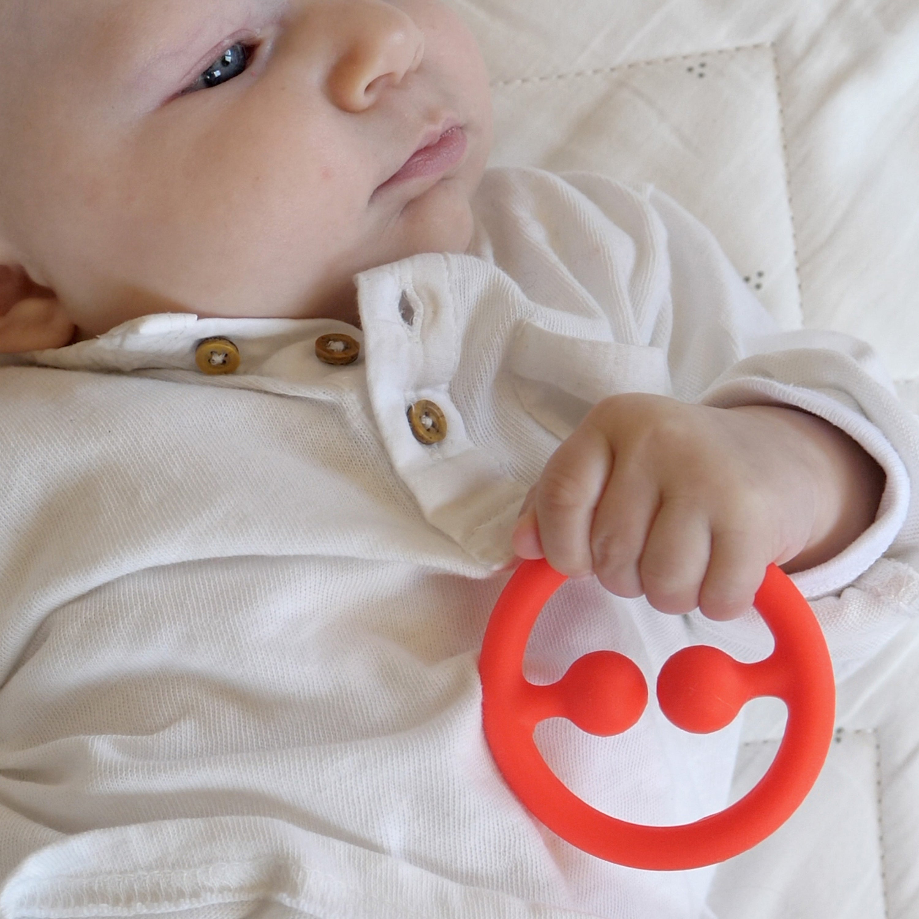 Baby holding a red Nigi Nagi Nogi silicone teething ring by MOLUK, a tactile toy for grasping and sensory learning.