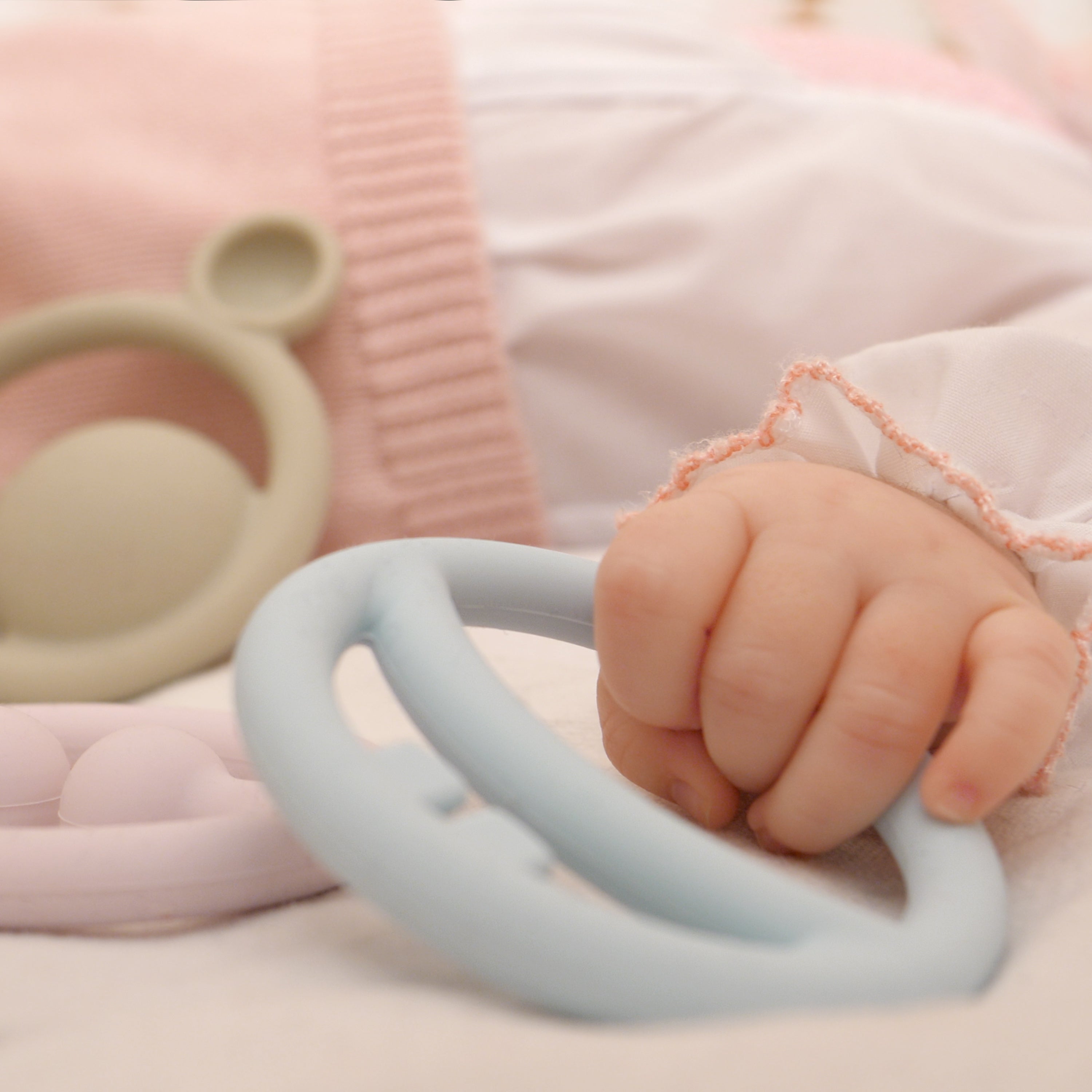 Baby’s hand holding an ice blue Nigi Nagi Nogi silicone teething ring by MOLUK, a tactile toy for grasping and soothing gums.