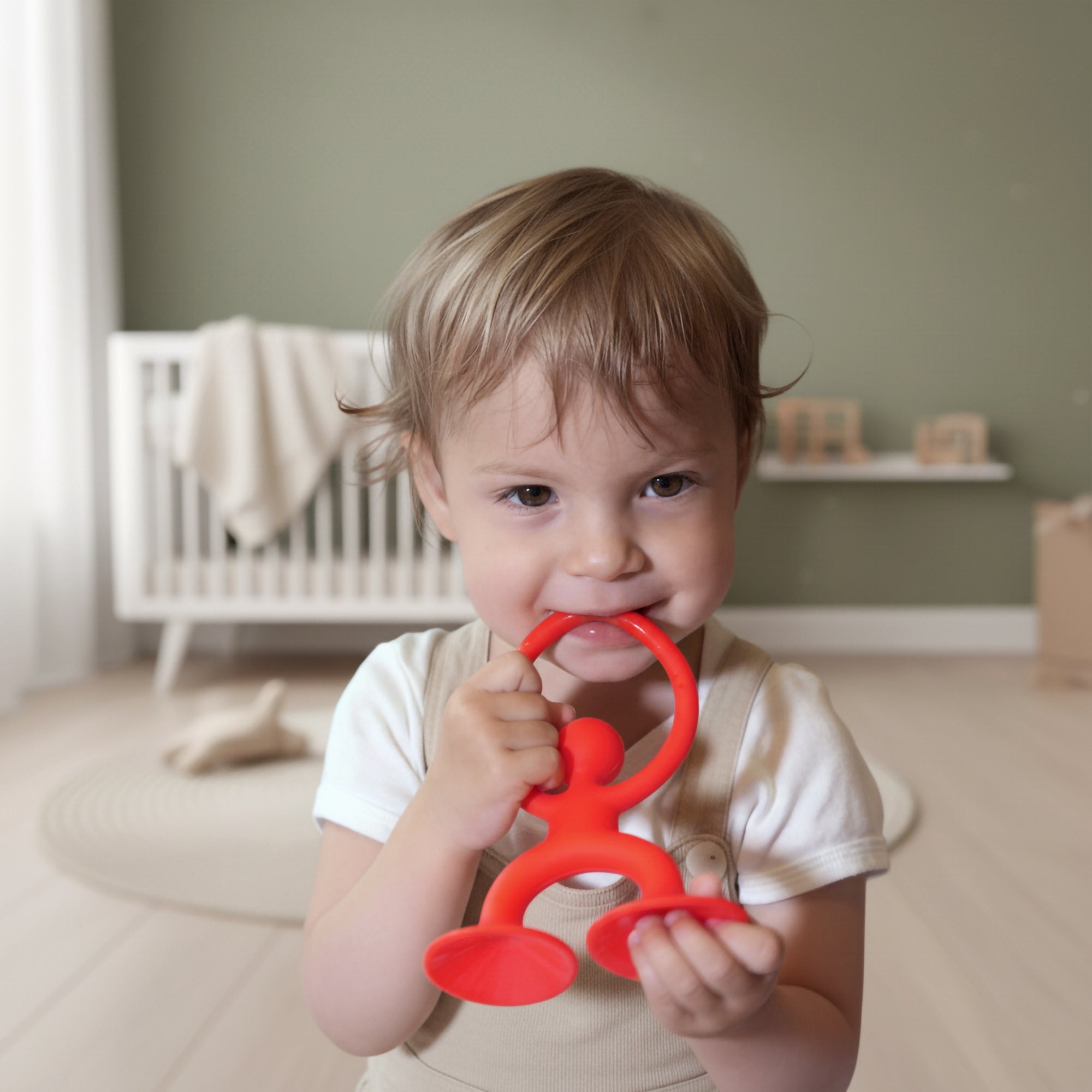 Toddler chewing on MOLUK’s red Noogi silicone teething and grasping toy in a modern nursery.