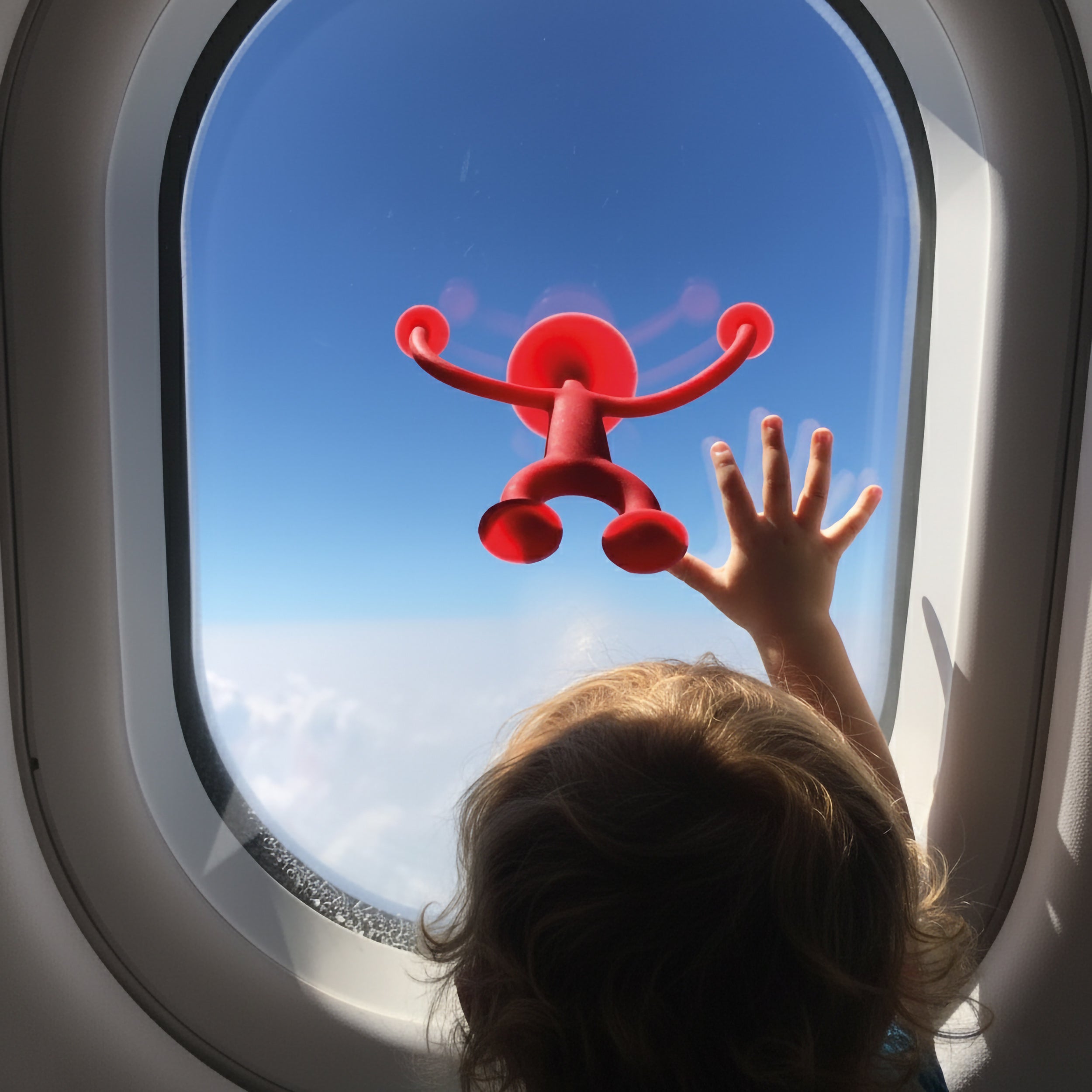 Toddler playing with a red Oogi suction cup silicone figure by MOLUK sticking to an airplane window while traveling.