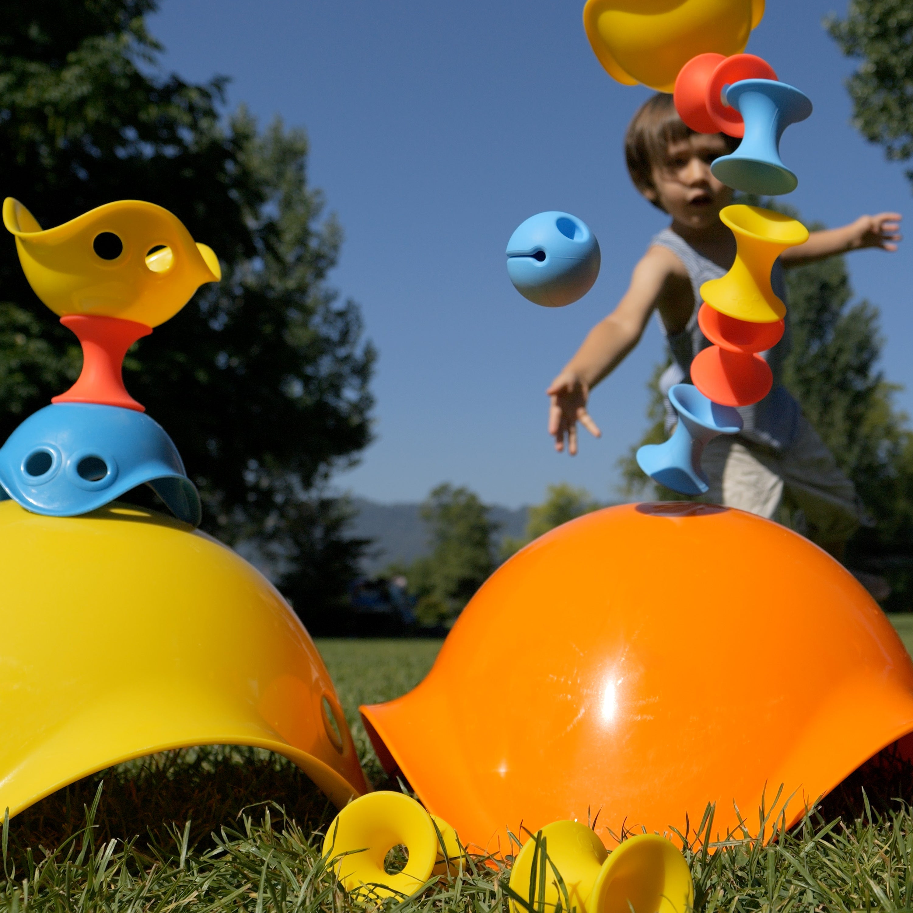 Child playing outdoors with MOLUK toys, tossing a blue Mox ball at a tower of Tulu silicone building blocks stacked on an orange Bilibo.