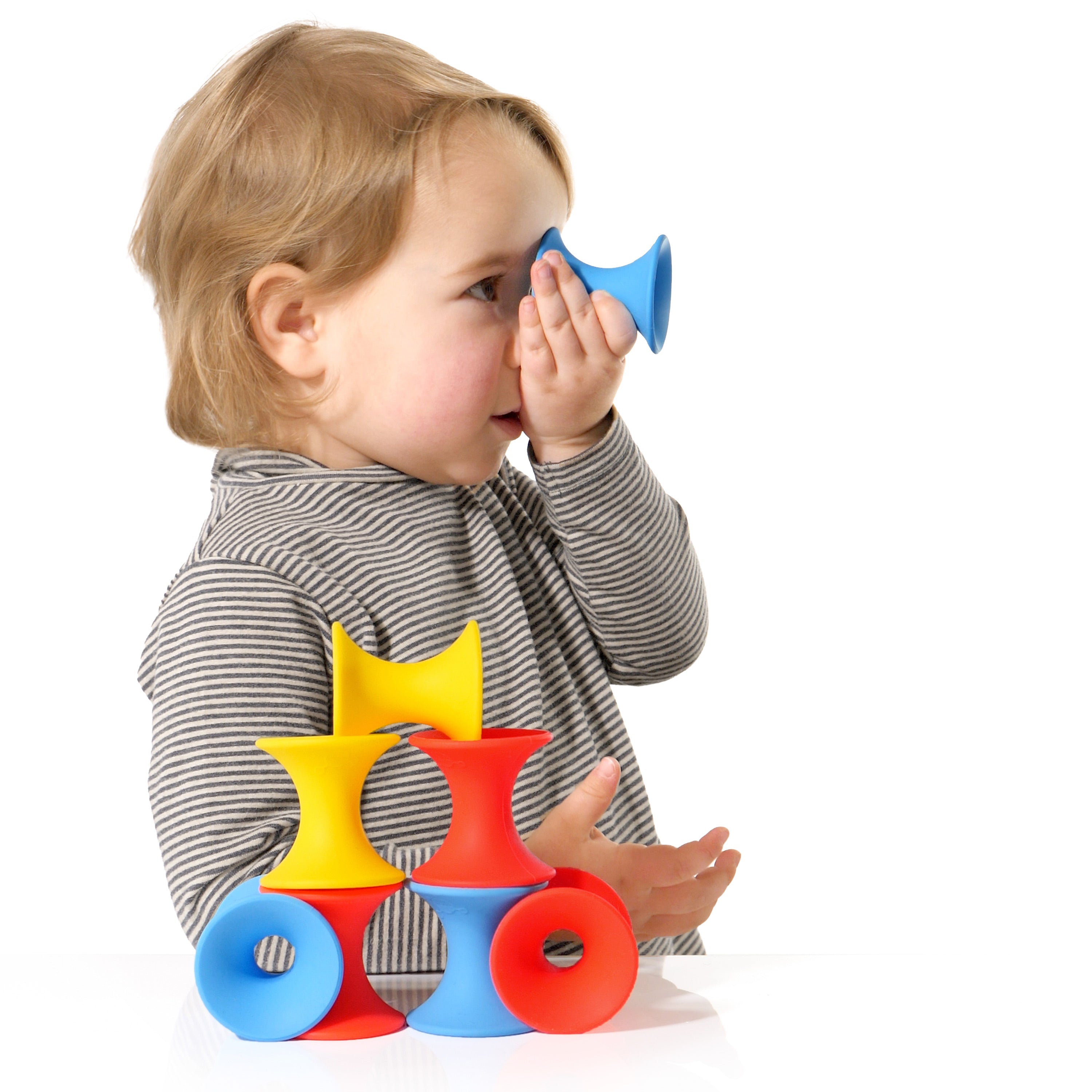 Child playing with MOLUK’s Tulu silicone building blocks, holding a blue piece to the eye like a telescope while colorful red, yellow, and blue pieces are stacked on the table.