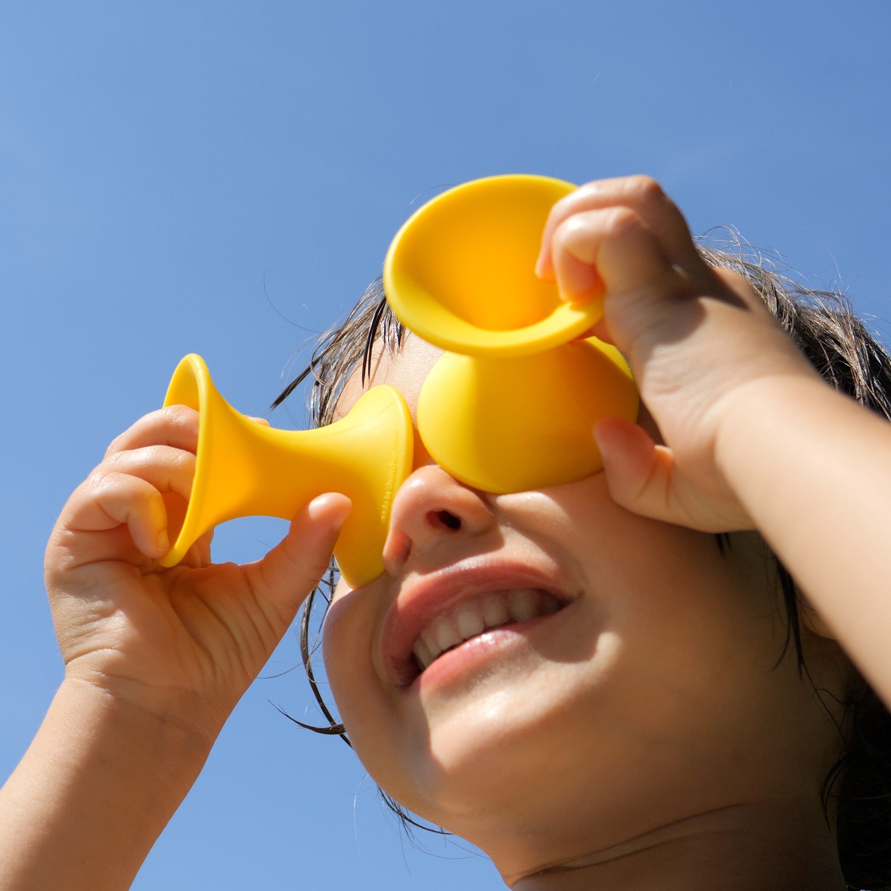 Child playing outdoors with MOLUK’s yellow Tulu silicone building blocks, holding them to the eyes like funny glasses under a blue sky.