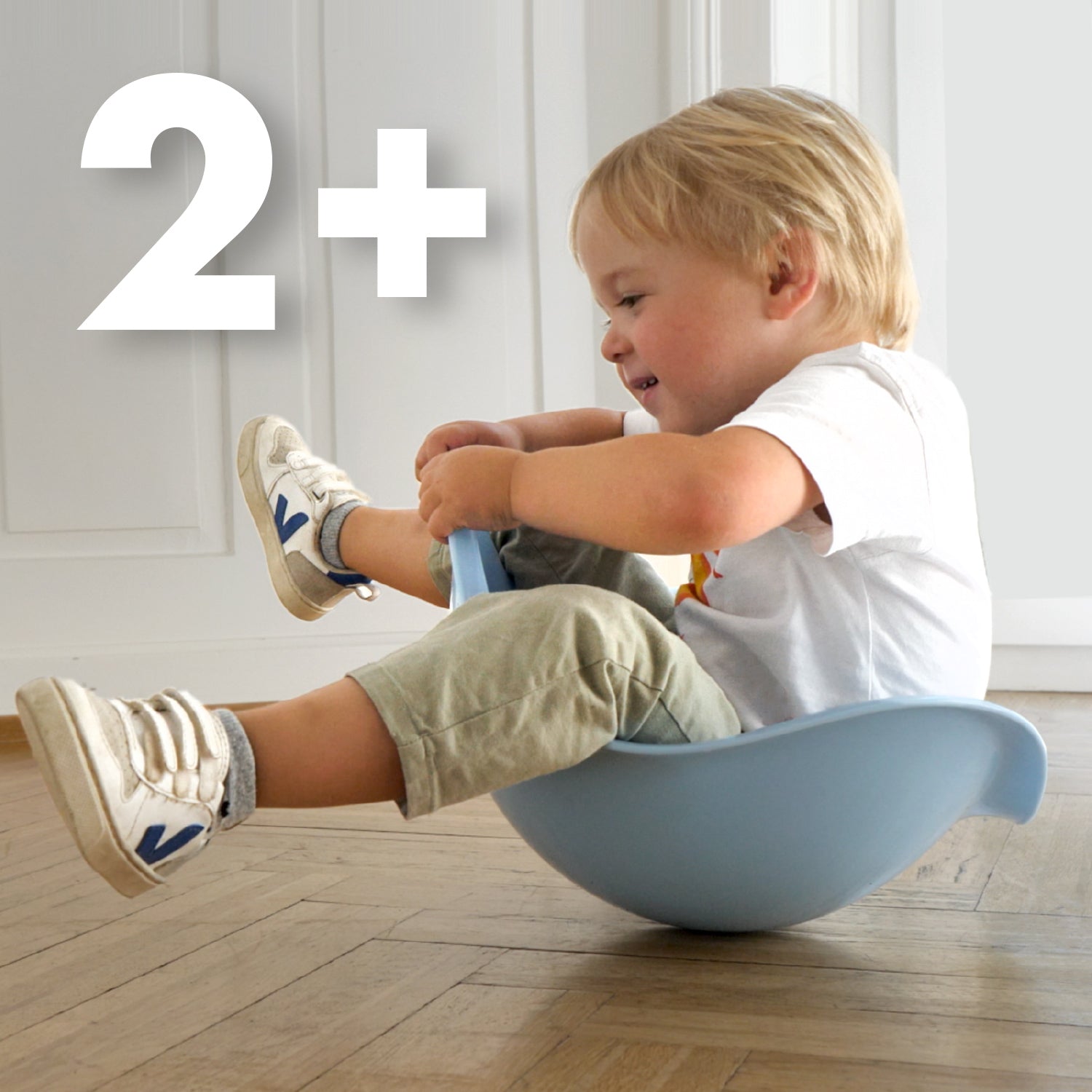 Child rocking inside  a blue Bilibo balance seat by MOLUK indoors with a '2+' age recommendation shown.