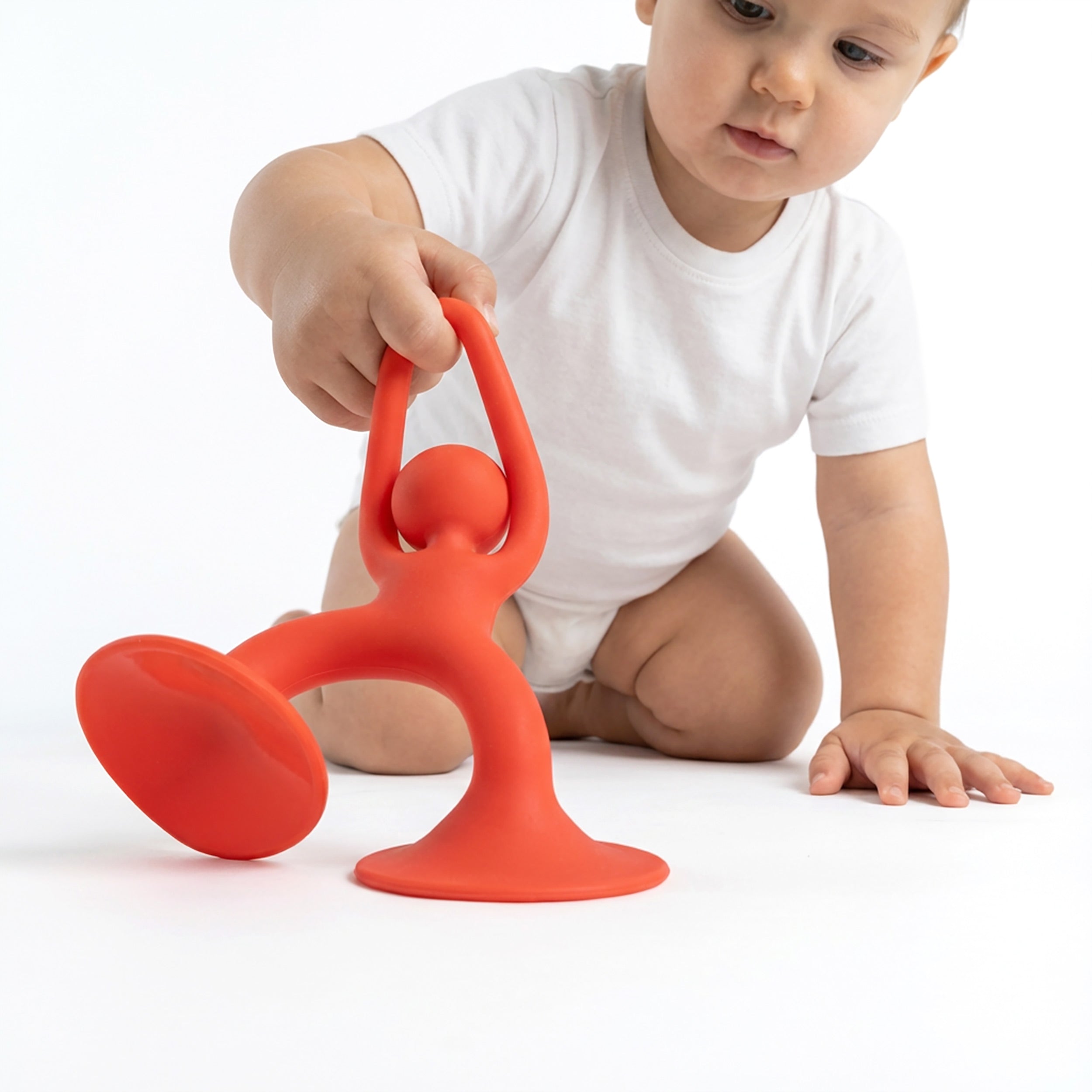Baby grasping bright red Noogi silicone toy by MOLUK, exploring its friendly shape and suction cup feet during floor play.